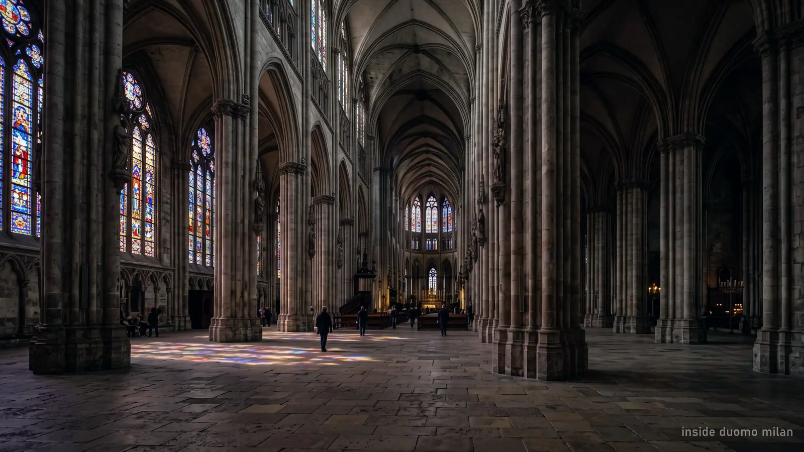 Inside a Gothic cathedral nave, clustered stone columns receding into shadow with coloured light from the stained glass
